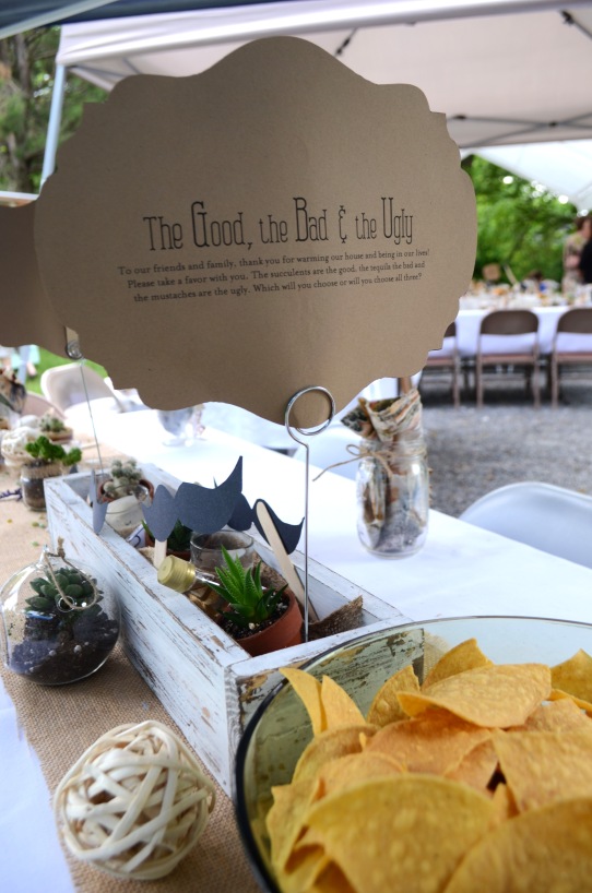 The sign explains how the favors are located in the middle of the table. It reads as follows: To our friends and family, thank you for warming our house and being in our lives! Please take a favor with you. The succulents are the good, the tequila the bad and the mustaches are the ugly. Which will you choose or will you choose all three? 